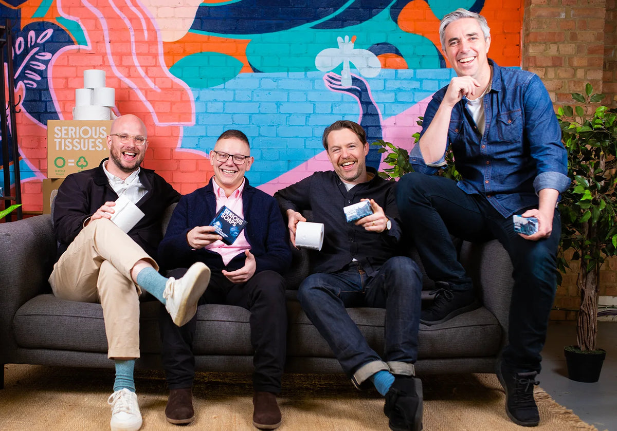 Four men sitting on a couch holding toilet paper in front of a colorful wall.